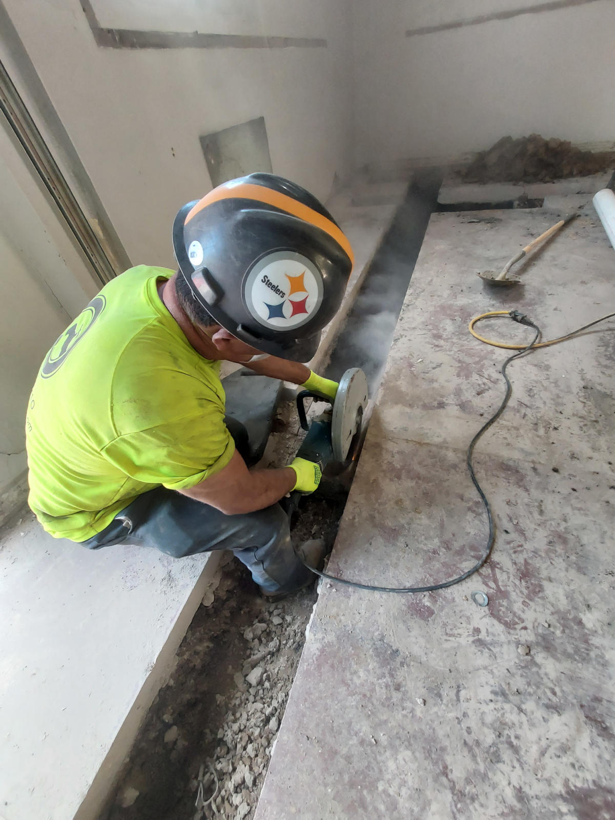 Plumber, who is Bill Helmken, using a concrete saw to cut slab for trench. Commercial plumbing Center for Disease Control. He is wearing a hard hat and kneeling down while using tool.